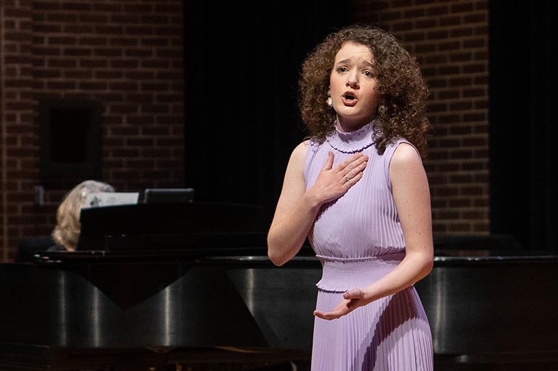 A student in a purple dress singing with someone playing piano behind her.