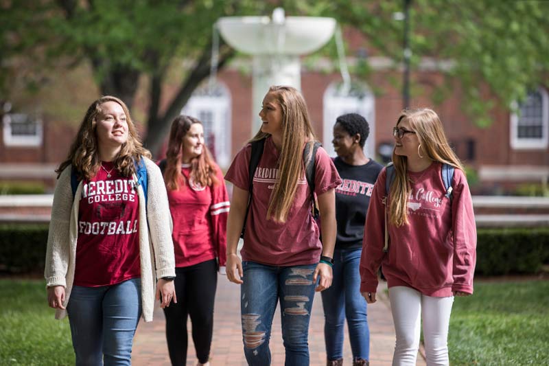 Five Students wearing black, maroon and white Meredith attire walking and talking in the campus quad with Heck Fountain and Belk Dining Hall visible.
