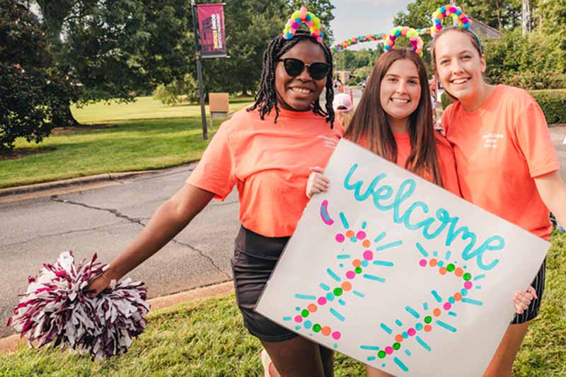 Three students at the entrance to Meredith on move-in day with pom-poms, balloon hats, and a sign that says "Welcome 2027".