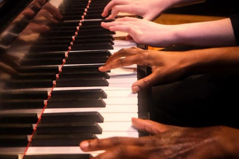 Two people, one with dark skin and one with light, playing the piano.