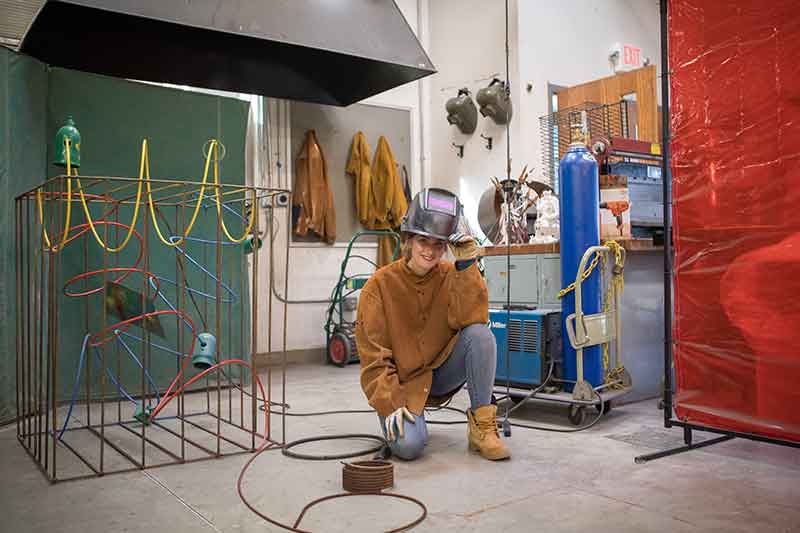 A student crouching and smiling as she lifts her welding mask, next to a sculpture she is creating.